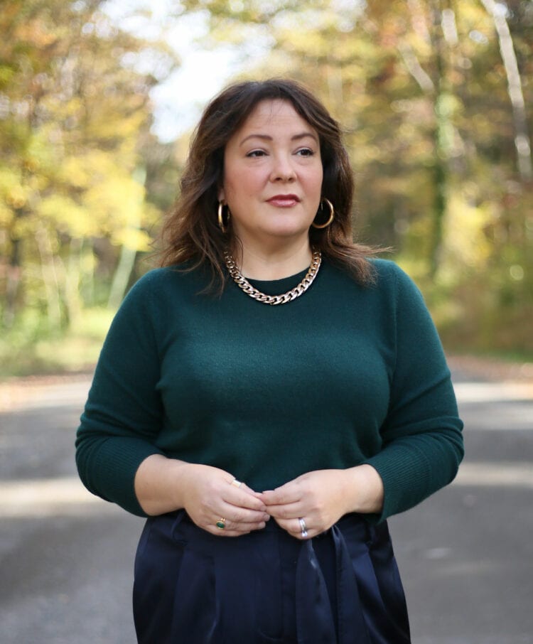 Alison is wearing a dark green Universal Standard crewneck sweater with a gold chain necklace and gold hoops. She is looking away from the camera with a thoughtful expression.