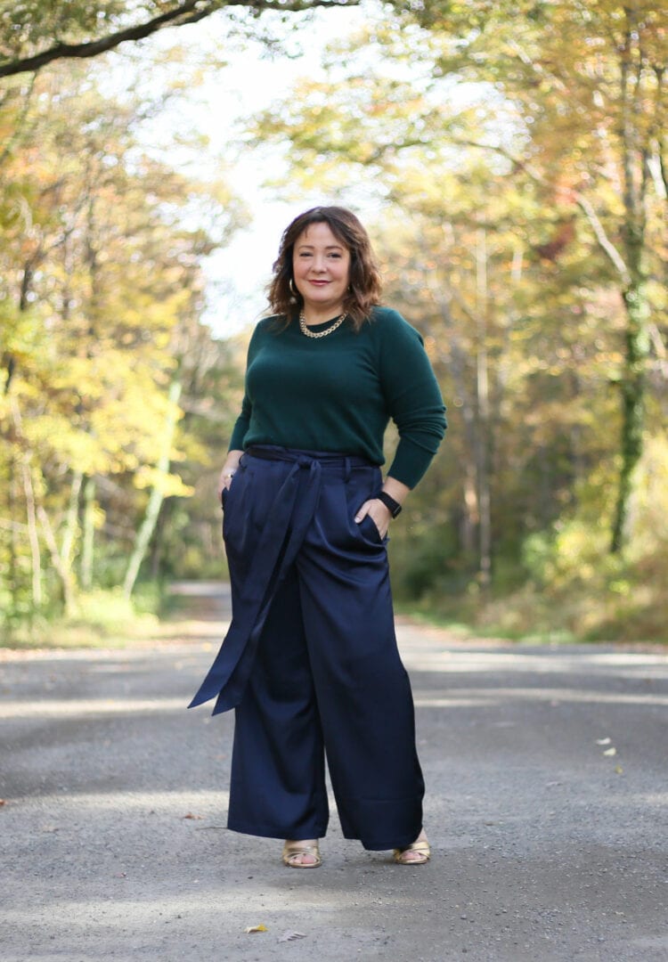 Alison of Wardrobe Oxygen is wearing a Universal Standard cashmere crewneck in dark green tucked into navy wide leg pants. Her hands are in her pockets and she is smiling at the camera. She is standing on a road going through a woods on a sunny fall day.