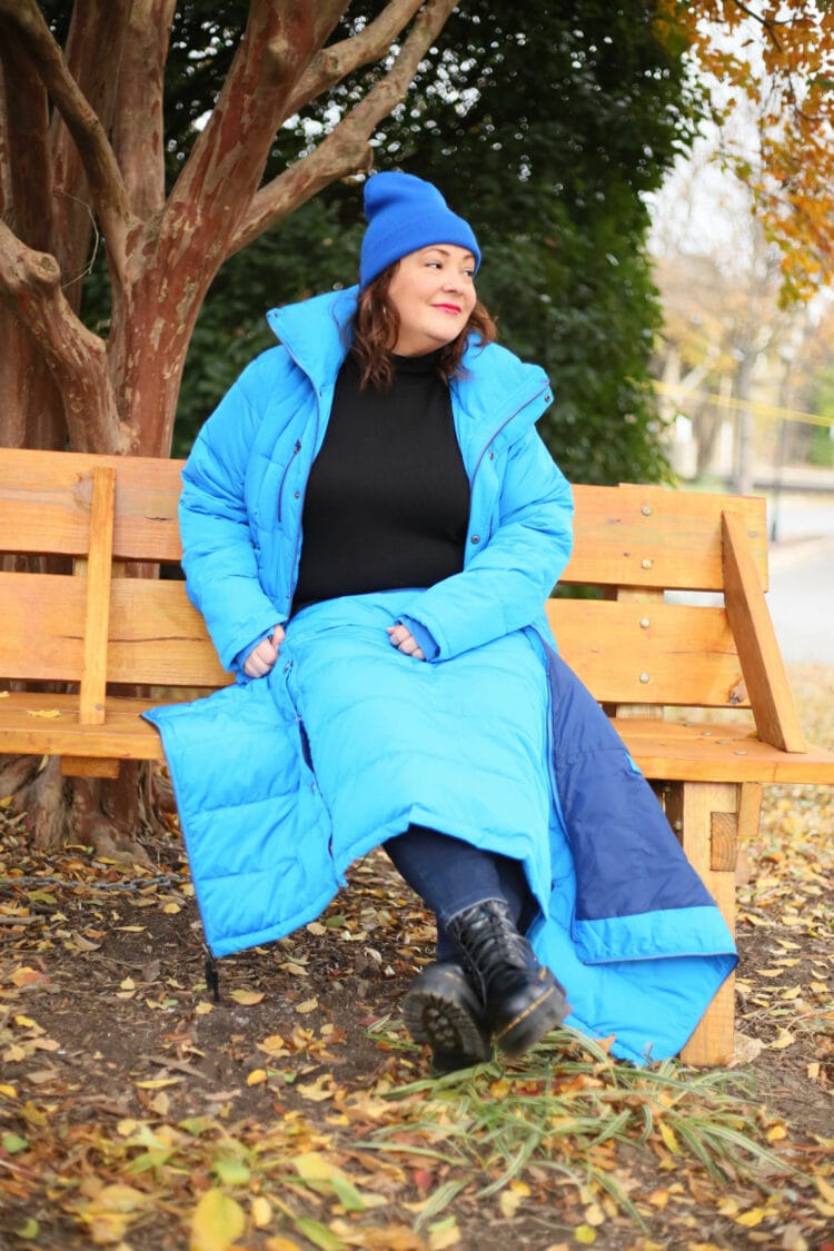 Alison in the short blue puffer coat sitting on a bench. She has taken the rectangle of the coat's skirt and placed it over her lap like aq blanket.