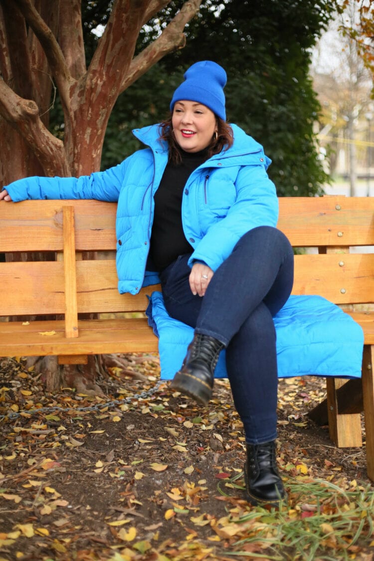 Alison is sitting on a bench with her legs crossed, her arm along the back of the bench. She is looking away from the camera, smiling. She is wearing the shorter version of the puffer, it is unzipped and you can see her black turtleneck sweater. She is sitting on the skirt of the coat; the fabric is folded in half to make a square pad on the bench.
