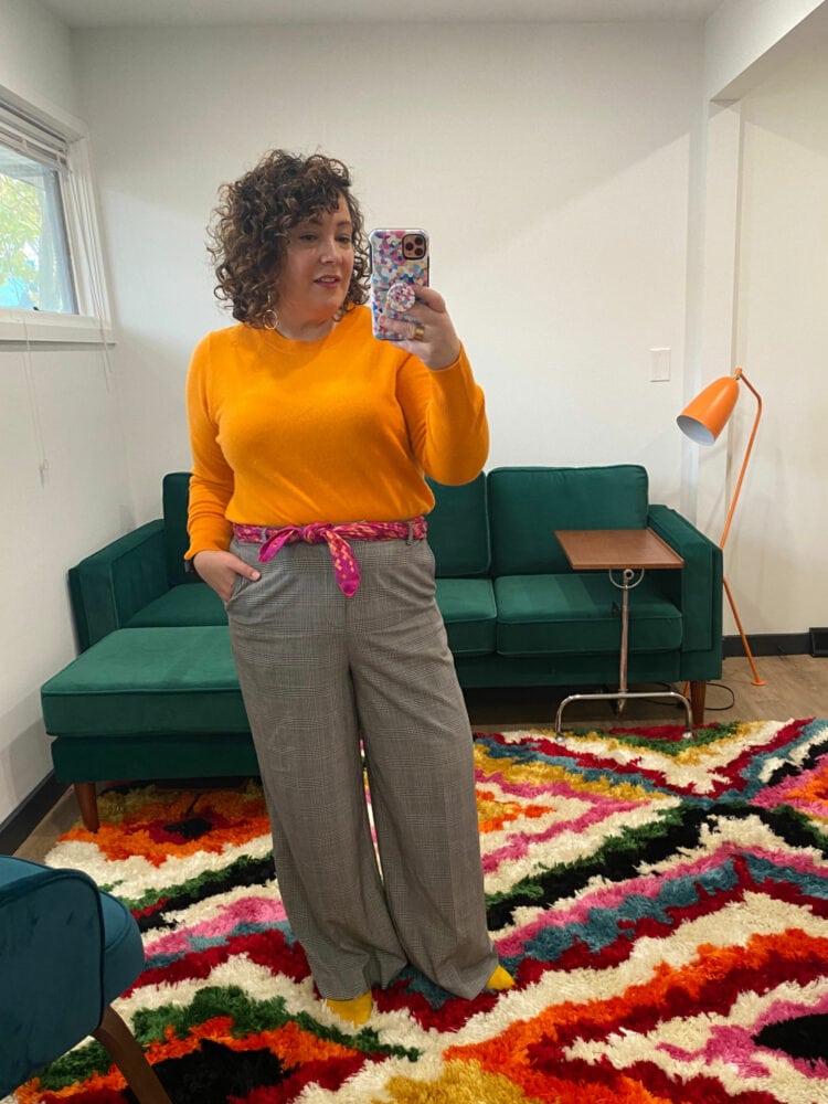 Alison is wearing a light orange sweater tucked into gray plaid wide leg pants with a pink scarf tied in a knot at her waist through the pant's beltloops.