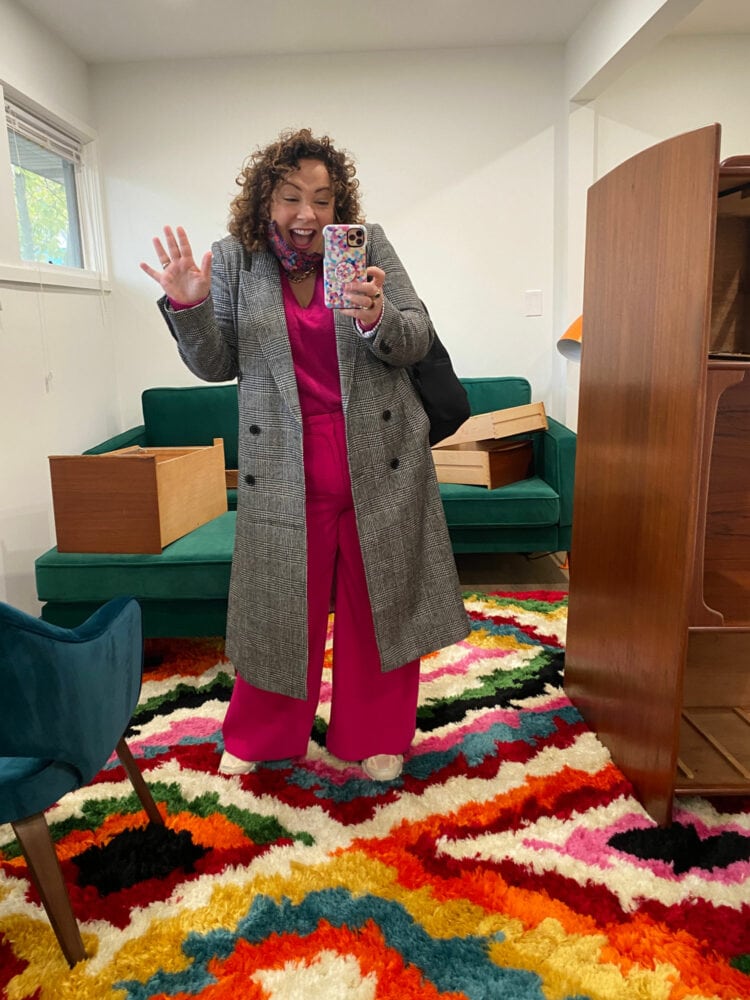 Alison in a gray plaid coat and pink pants looking excited and taking a selfie in the mirror of her office