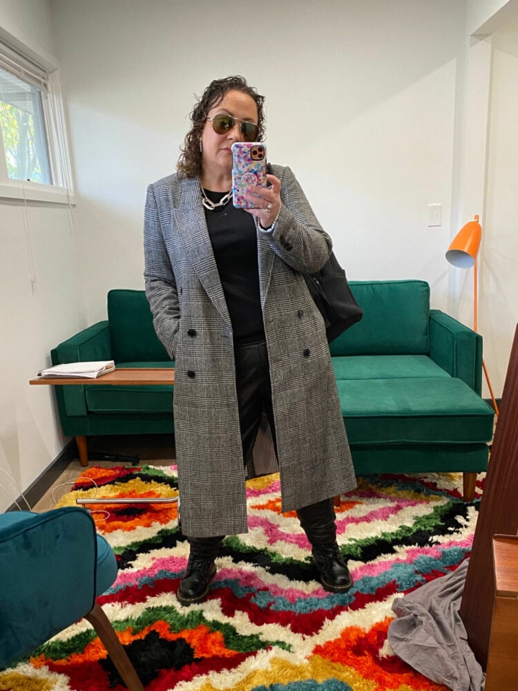 Alison taking a mirror selfie in her office wearing all black with a gray plaid long coat and aviator sunglasses.