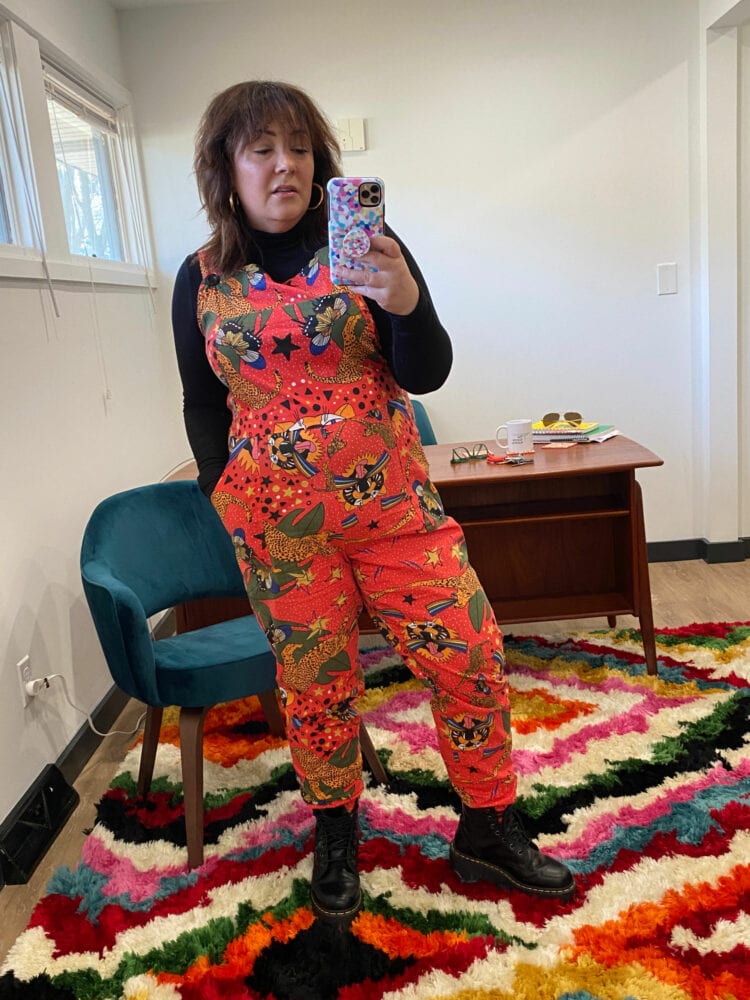 Alison wearing orange printed cotton overalls from the brand Nooworks in the print Tiger Pride. She is wearing them over a black fitted turtleneck with black Dr. Martens boots. She is standing in front of a Mid Century Modern desk and teal velvet chair
