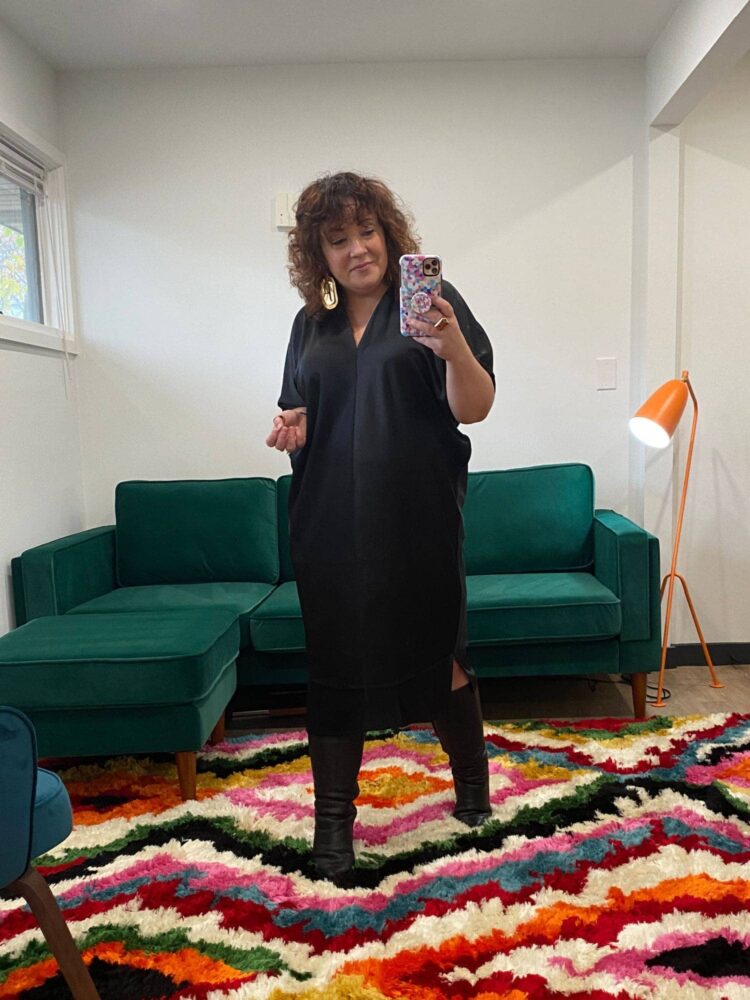 Alison wearing a black satin v-neck calf length short sleeved caftan from Universal Standard with heeled knee high boots. She is doing a mirror selfie in front of a green velvet sofa from Albany Park.