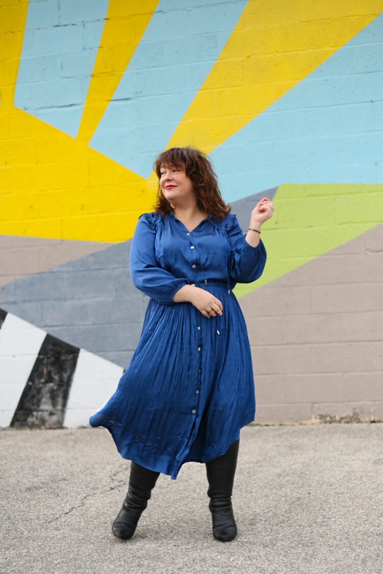 Alison wearing a blue silky drawstring waist longsleeved dress with heeled black knee high boots. She is standing in front of a graphic mural and the wind is catching the hem of her skirt.