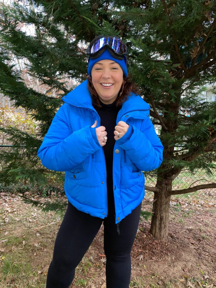 how to dress aprés ski: Alison is wearing an electric blue puffer coat and matching knit beanie with a black turtleneck catsuit underneath. She is smiling at the camera, holding her jacket closed and she has blue and black ski goggles on the top of her head over the beanie.