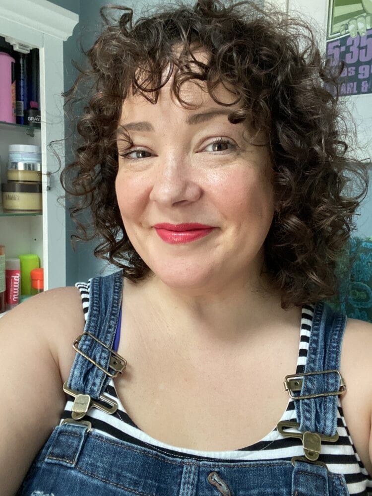 Alison in the summer with curly hair. She is wearing a black and white striped tank top with overalls and bright pinkish red lipstick. It is a selfie, she is holding the phone and smiling at the camera.