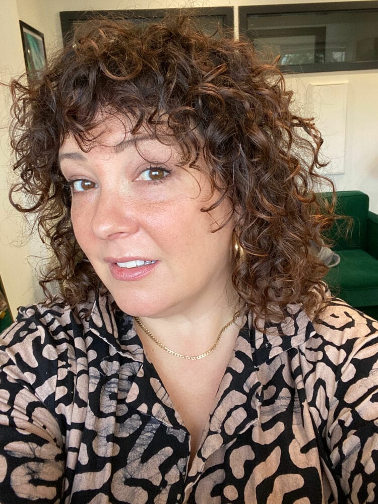 Alison looking at the camera with curly hair. She is wearing a tan and black graphic print cotton jumpsuit with gold hoops and a gold cuban link chain necklace.