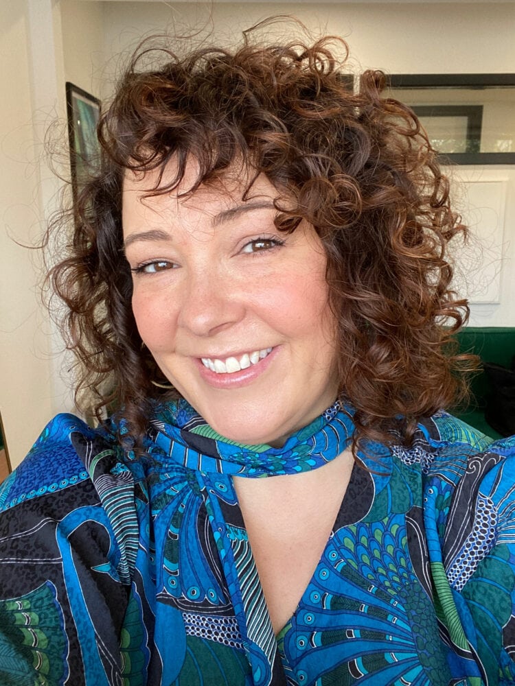 Alison with curly hair smiling at the camera. She is wearing a blue paisley silky blouse with a scarf neckline.