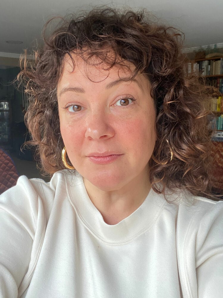 Alison is doing a camera selfie, looking at the camera. She has fluffy undefined curls and is wearing a cream neoprene sweatshirt and gold hoops.