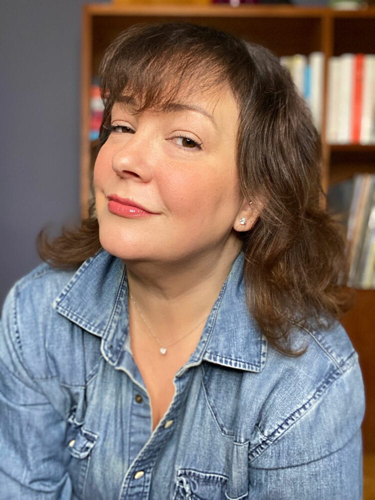 Alison Gary of Wardrobe Oxygen in a denim shirt looking at the camera and smirking. She is wearing James Allen diamond studs in her ears and a delicate silver chain with a diamond pendant on it.