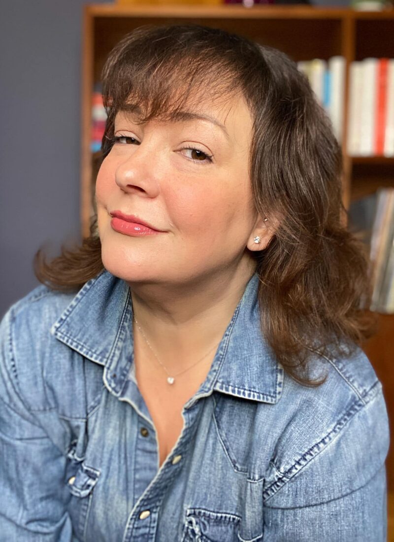 Alison Gary of Wardrobe Oxygen in a denim shirt looking at the camera and smirking. She is wearing James Allen diamond studs in her ears and a delicate silver chain with a diamond pendant on it.