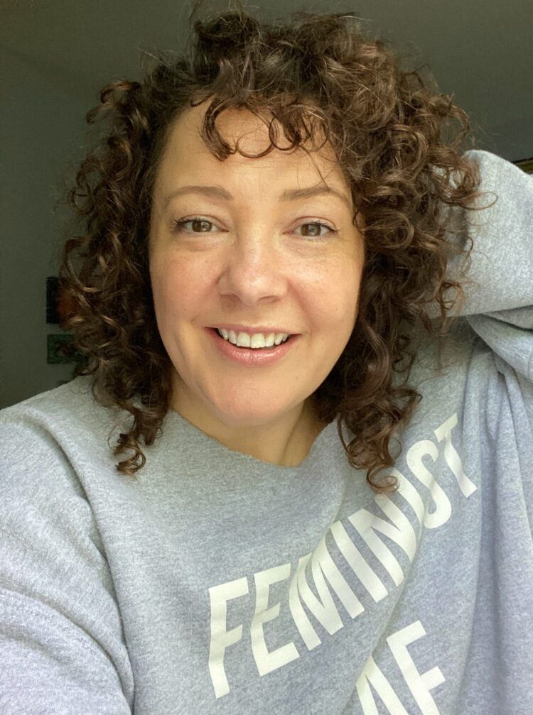 Alison Gary of Wardrobe Oxygen smiling at the camera, not wearing makeup. She is wearing a gray sweatshirt that says Feminist on the front