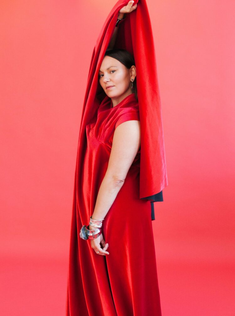 Image of Tanya Tagaq, Canadian musical artist. Tagaq is standing in front of a dark pink background. She is wearing a long red satin sleeveless shift dress and is holding the train of the dress over her head to almost create a hood. She is facing to the left of the camera but looking directly into the camera.