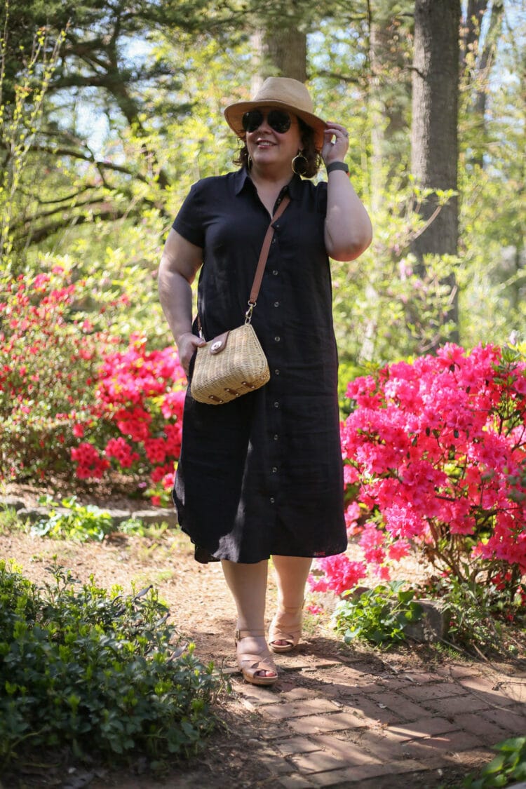 Alison in the Universal Standard Dune linen shirtdress in black. She is wearing a straw fedora and wicker crossbody bag and black sunglasses and is walking down a path bordered with azalea bushes in bloom