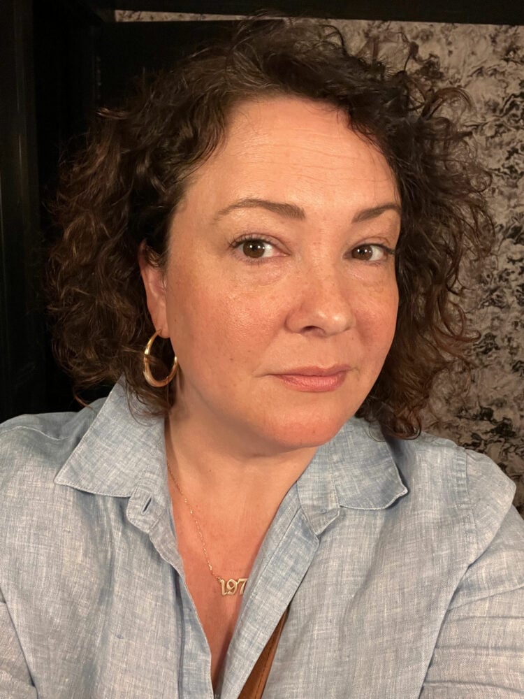 Selfie of Alison Gary wearing a blue linen shirt, gold hoop earrings and a gold necklace with a "1975" charm. She has shoulder length curly brown hair and is looking at the camera
