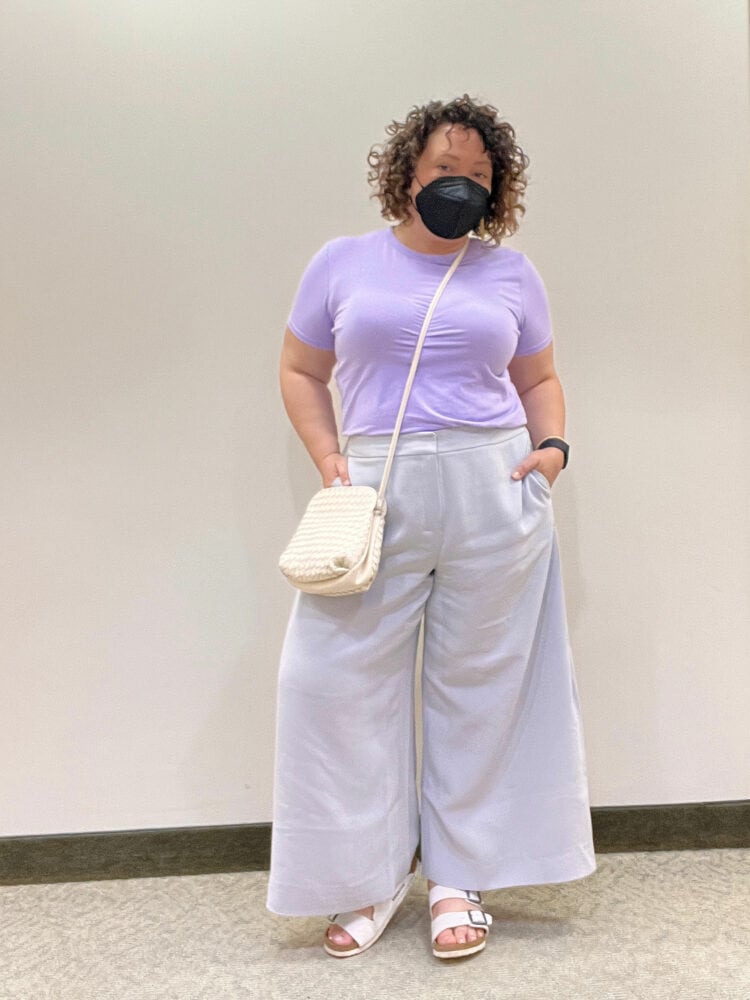 Photo of Alison Gary of Wardrobe Oxygen. She has chin-length curly brown hair and is wearing a lavender short-sleeved t-shirt tucked into pale gray crepe ankle-length wide leg trousers. She has on white Birkenstock sandals and a cream woven leather crossbody bag.