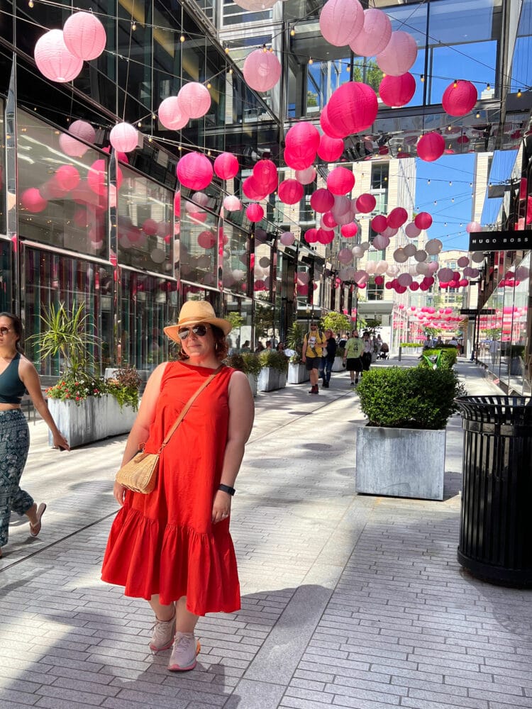 A woman in an orange cotton sleeveless dress and straw hat standing in City Center DC, a line of luxury shops and the  outdoor space decorated with paper lanterns in shades of pink.