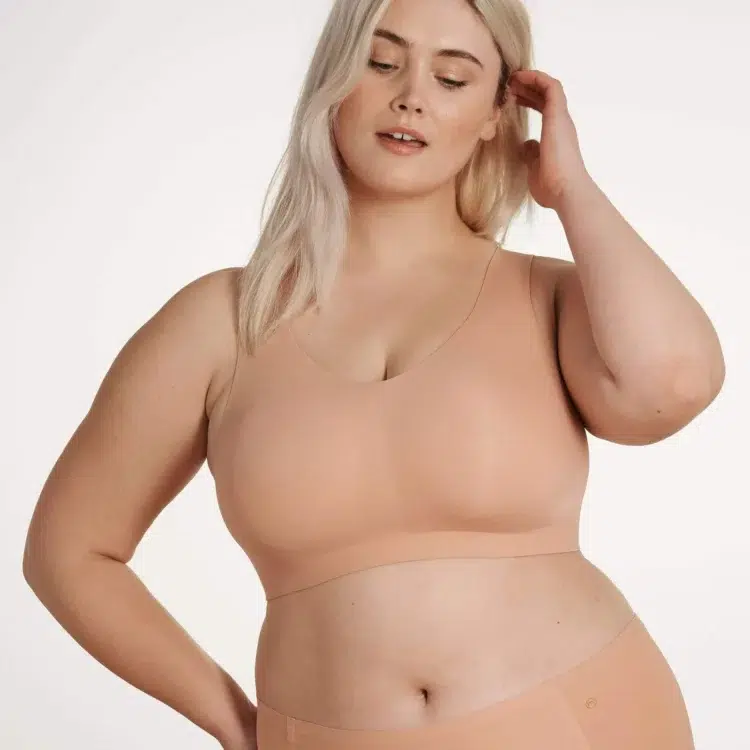 A blonde curvy model wearing the Evelyn & Bobbie Defy bra in a beige color that matches her skin