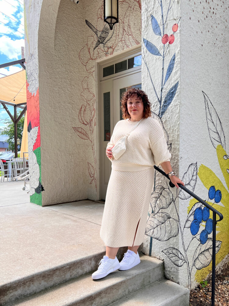 Alison is leaning against the muraled wall at the front of Pennyroyal Station, a restaurant on busy Rhode Island Avenue in Mount Rainier, a city on the Washington DC border. She is wearing a cream sweater knit calf-length skirt and matching crewneck with white Adidas sneakers. She has a Brandon Blackwood cream tumbled leather belt bag worn as a crossbody; she is touching the bag and smiling at the camera.
