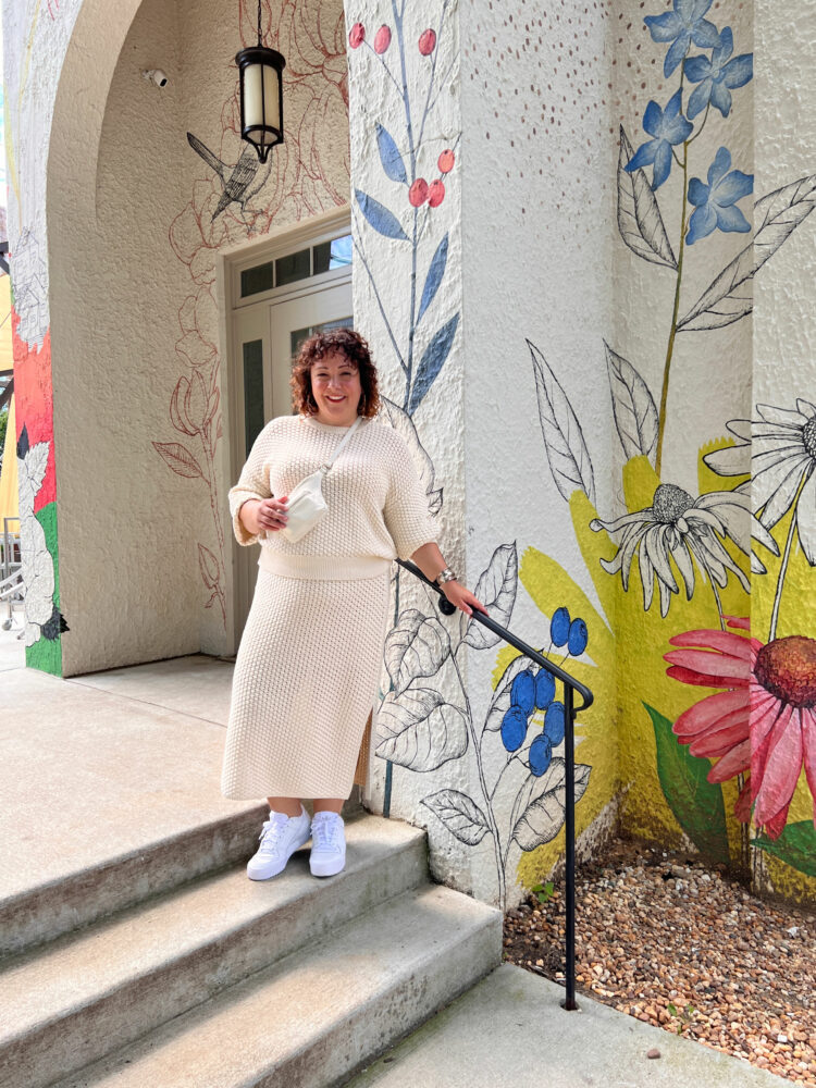 Alison Gary of Wardrobe Oxygen outside Pennyroyal Station restaurant in Mount Rainier, Maryland. She is wearing a cream popcorn knit M.M.LaFleur sweater set of calf-length skirt and matching long-sleeved crewneck. She has a cream leather belt bag as a crossbody and is wearing white Adidas sneakers.