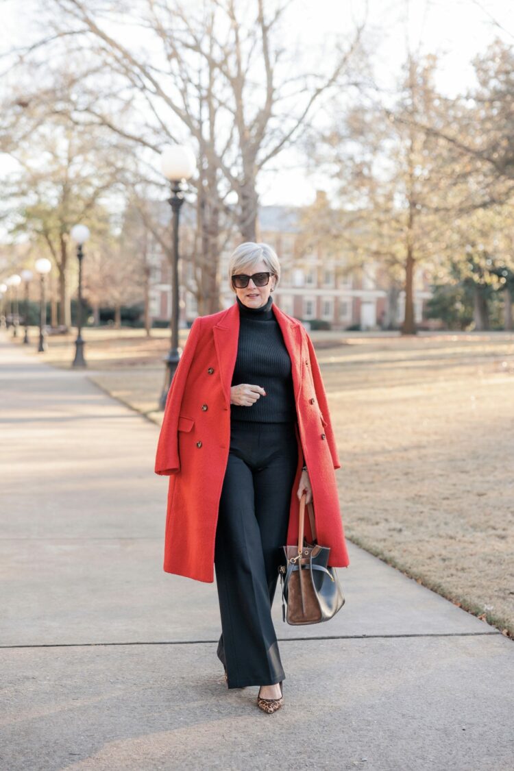 Influencer and over 50 blogger Beth Djali from Style at a Certain Age styles a black turtleneck and trousers with a red wool double breasted winter coat and leopard print kitten heel pumps. 
