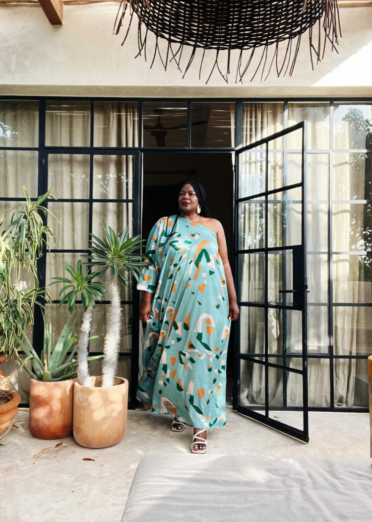 Influencer Assa Cisse from the blog My Curves and Curls wears a pale aqua printed one shoulder maxi length caftan from the brand diarrablu