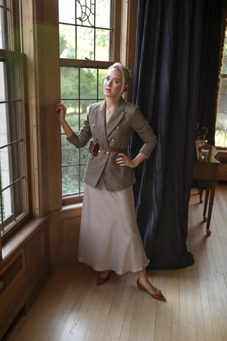 Mary Orton, influencer also known as Memorandum. Orton is wearing a double breasted khaki tweed blazer with a beige silk slip-style skirt. She has belted the blazer with a brown leather belt bag and on her feet are brown pointed toe flats.