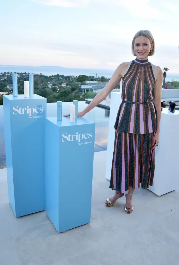 Naomi Watts in a cocktail dress on a rooftop patio standing with a display of her menopause products called Stripes