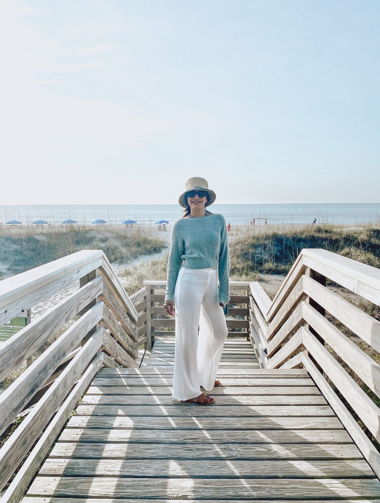 Influencer Sapna Delacourt who is also known from her blog and social media platforms Lunch with a Girlfriend is wearing a pale blue linen sweater and white lightweight beach pants with a straw bucket hat and brown leather fisherman sandals. She is standing on a wood path to a beach, the ocean in the distance.