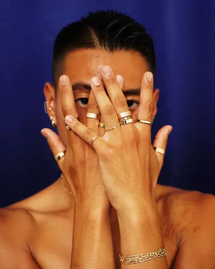 Image of a person wearing at least one gold ring on every finger; their hands are in front of their face and they are peeking through their fingers. The rings are from Automic Gold a company that offers stylish rings for larger fingers.