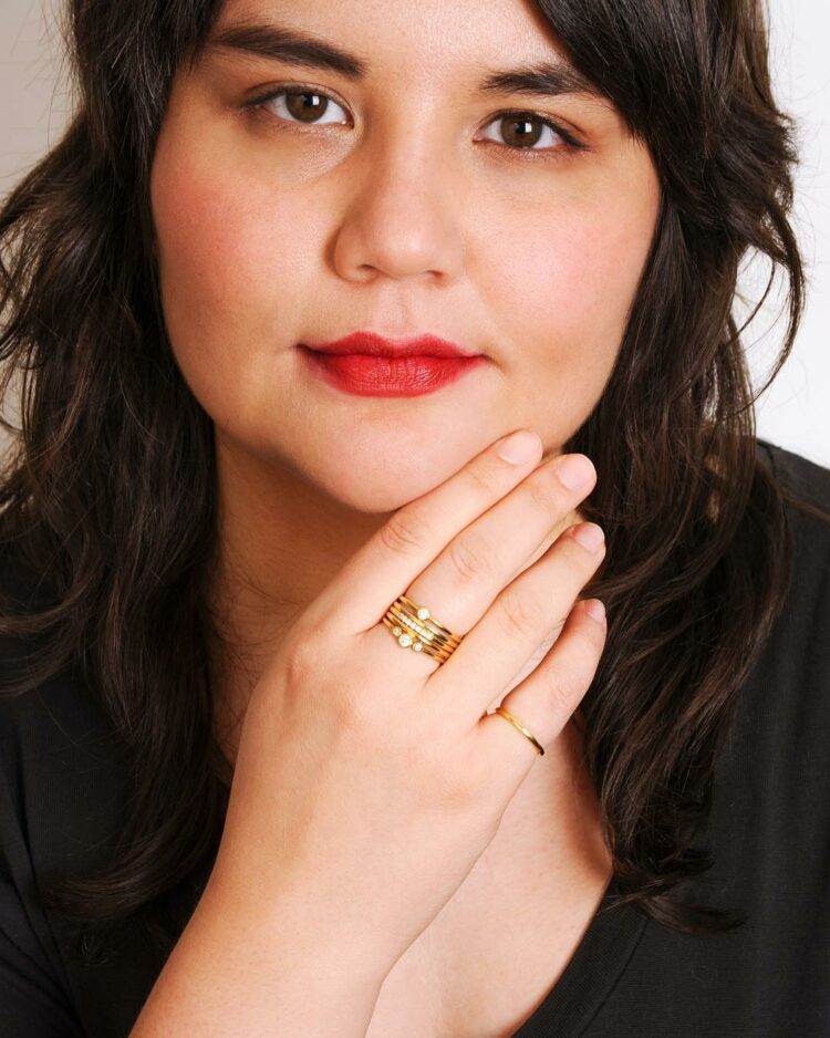 Image of a woman with dark hair and red lips holding up her hand which features multiple gold rings from the brand Poirier, which carries demi fine stylish rings for larger fingers.