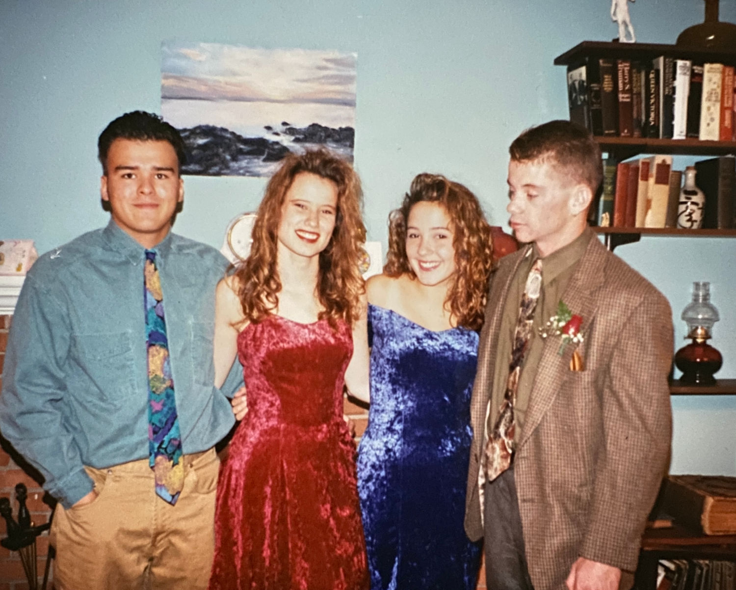 Four teenagers in 1992. The two girls are wearing crushed velvet homecoming dresses. The boys are wearing colored button-down shirts with khaki pants and floral print ties. They are standing in a living room in front of a fireplace flanked by bookcases.