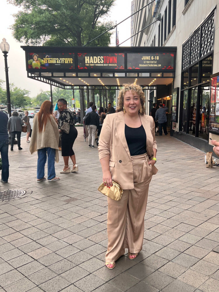 Alison Gary of Wardrobe Oxygen in front of National Theatre in Washington DC. She is wearing a tan satin pantsuit with a black shell underneath and gold heels. She is holding a gold clutch in her hand.