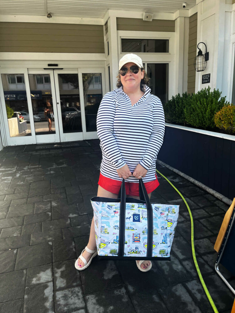 Alison Gary of Wardrobe Oxygen at the Avenue Inn in Rehoboth Beach, Delaware. She is wearing a navy and white striped zip front tunic rash guard from Lands' End with red Sunny shorts from Universal Standard. She is carrying a Scout Deano bag with Washington DC icons on it and on her feet are Teva Hurricane Drift sandals.