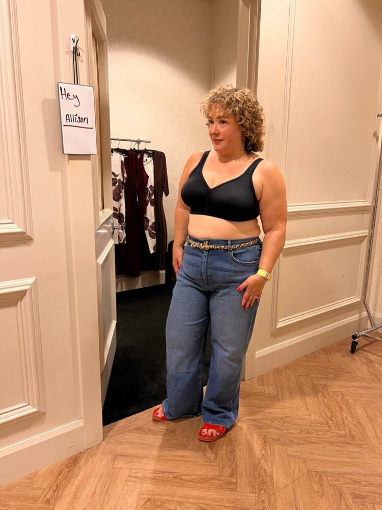 Alison looking at her reflection in a Soma Intimates fitting room wearing a black wireless bra and jeans