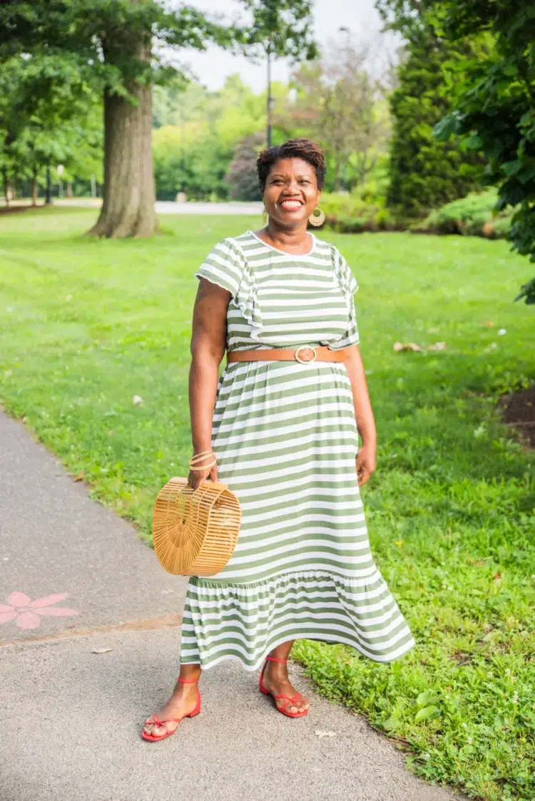 Photo of Georgette Niles, an over 40 fashion blogger and influencer. She is wearing a sage green and white striped sundress with a wicker purse, tan leather belt, and orange strappy flat sandals.