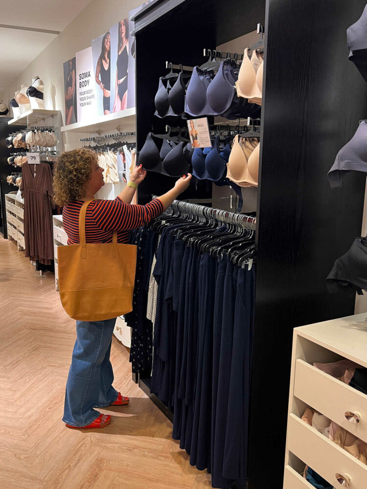 Alison of Wardrobe Oxygen in a Soma Intimates boutique. She is examining a bra that is hanging on a wall in a display.