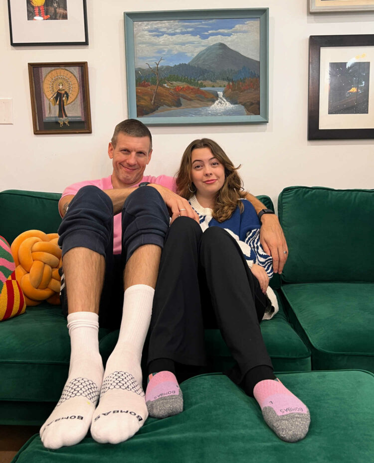My husband and teenage daughter sitting on a couch in loungewear. My husband is wearing the Bombas Original Crew socks in white, my daughter a pair of Bombas Ankle Running Socks in a colorway of pink, lavender, gray, and black.