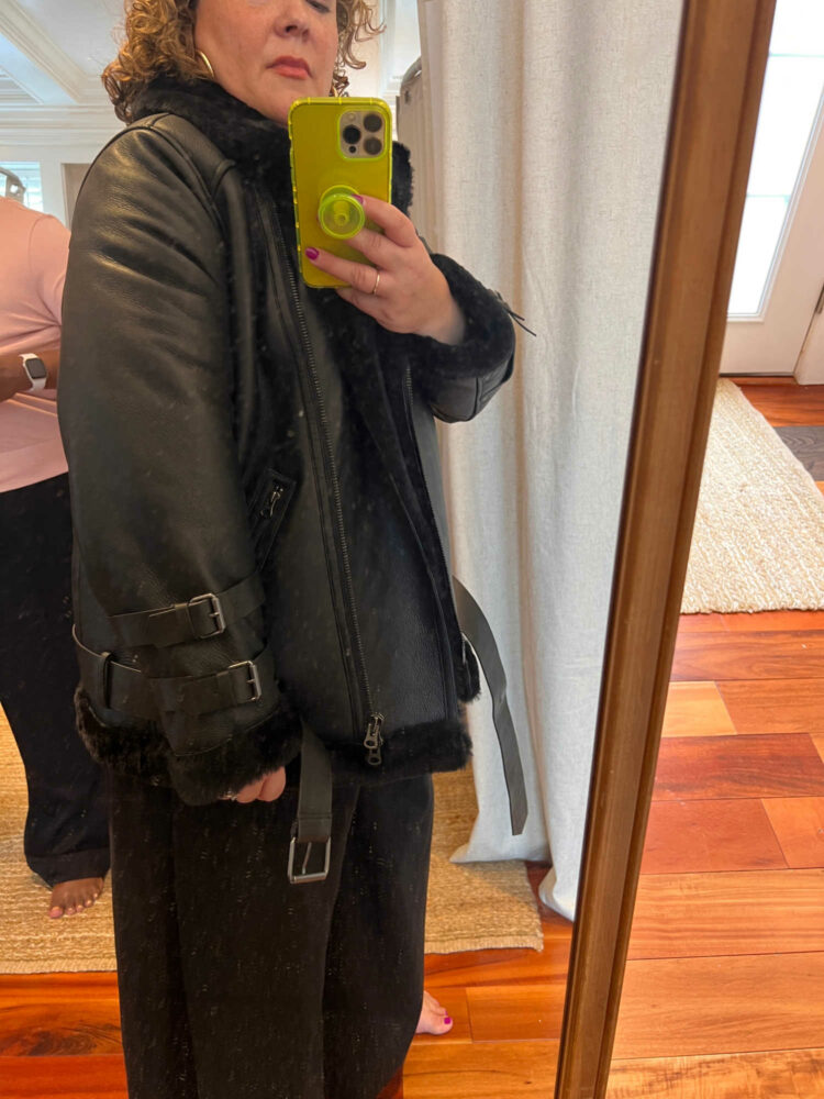 a woman trying on a leather and shearling coat and taking a picture of herself in a mirror with her iphone