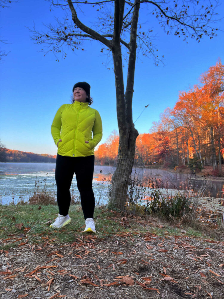 Alison in a highlighter yellow puffer vest, zip shirt, and black leggings standing in front of a lake.