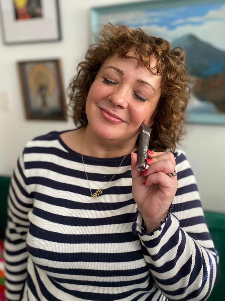 Alison Gary of Wardrobe Oxygen in a Breton striped top smiling and holing a Colleen Rothschild lip balm near her face, smiling
