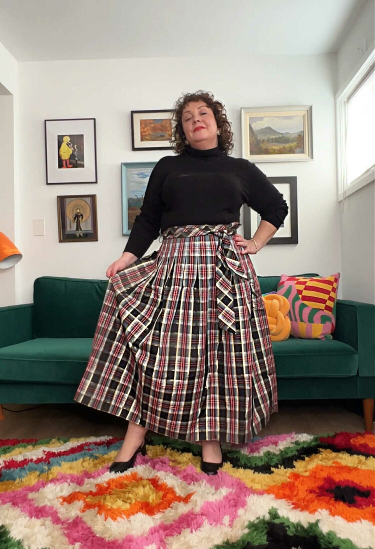 Woman with short curly brown hair wearing a black turtleneck sweater tucked into an ankle length full skirt of a metallic holiday plaid.