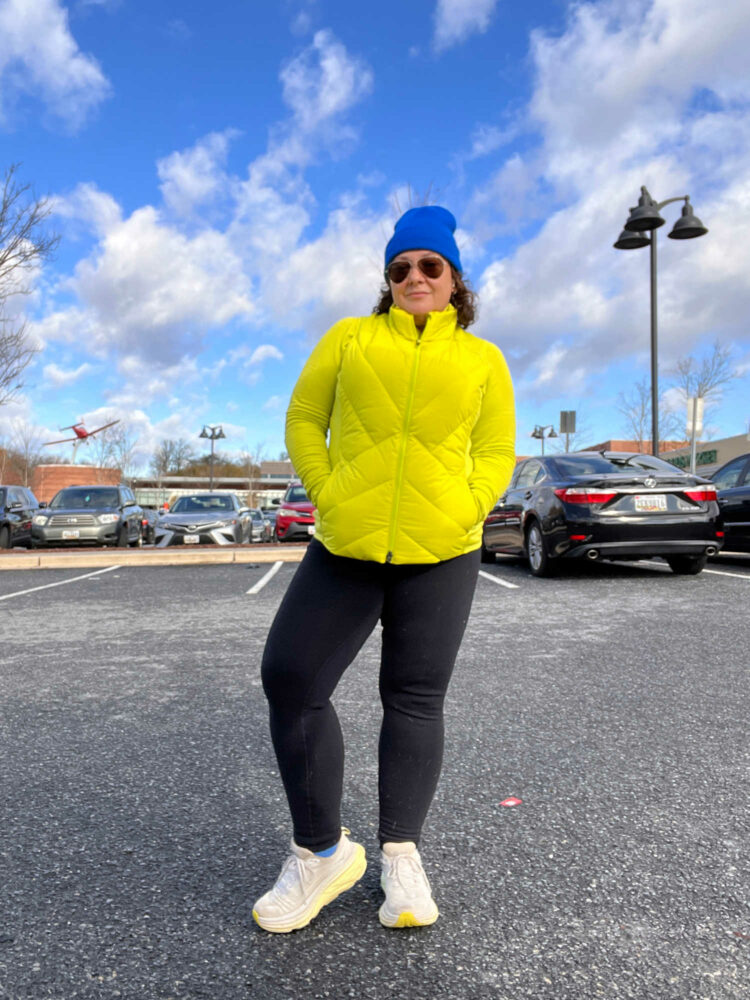 Woman standing in a parking lot wearing the Athleta Dark Fiber Optic vest over a half-zip long sleeved top. It is paired with black leggings, HOKA sneakers, and a bright blue knit beanie.