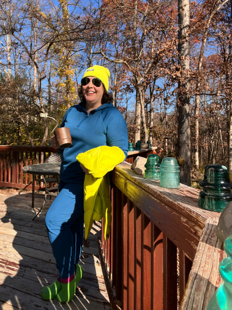 Alison in Athleta Dark Lapis Blue hoodie and joggers. She is standing on a deck wearing a Dark Fiber Optic beanie cap and holding a vest of the same color in her hand. She is laughing and holding a brown stonewear coffee mug. On her feet are green Birki clogs and hot pink running socks from Bombas