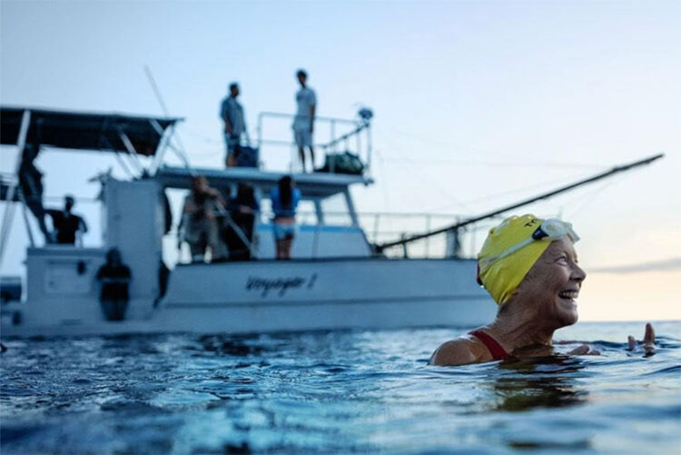 Scene from the movie Nyad of Annette Benning as Diana Nyad in a yellow swim cap smiling as she floats in the ocean, with a white boat in the background