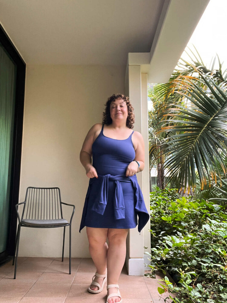 Alison Gary of Wardrobe Oxygen in the cobalt Universal Standard Next to Naked Dress with the coordinating Next to Naked jacket tid around her waist. She is standing on a patio of a hotel in a tropical locale, smirking at the camera while chinching the jacket around her waist.