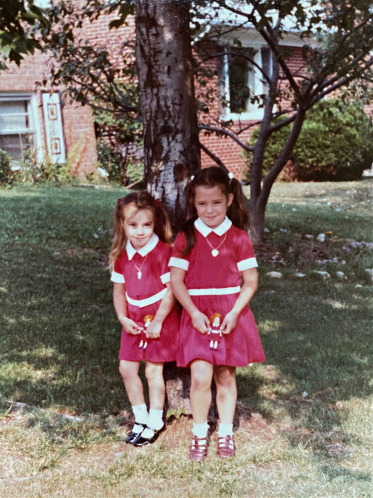 two young girls standing against a tree in the front yard of a suburban home in 1982. They are both wearing red dresses with white collar and cuffs, white ankle socks, and black patnet leather Mary Janes. They both have gold locket necklaces that were costume jewelry to promote the 1982 film Annie.