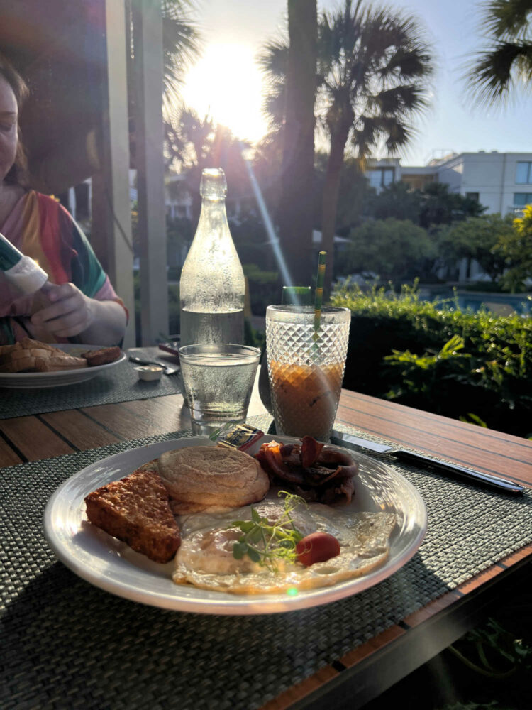 a plate of breakfast food and an iced coffee on an outdoor table overlooking tropical plants and a pool